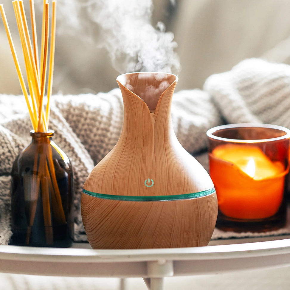 A wooden-look essential oil diffuser with misting at the top, styled on a white tray next to a reed diffuser and a glowing orange candle.